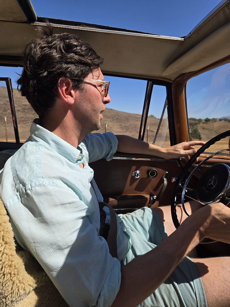 Man driving a classic car on a sunny day through scenic countryside with clear blue skies.
