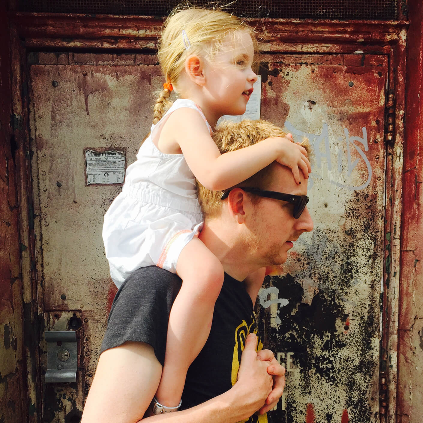 Man carrying child on shoulders in front of rustic door, bonding and sunlight play, summer adventure scene.