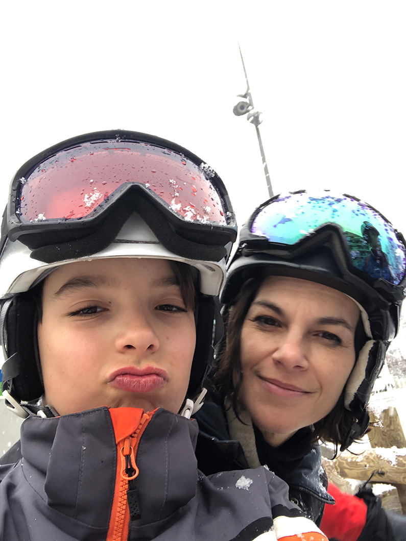 Mother and child on a ski trip, both wearing helmets and goggles, with snowy background.
