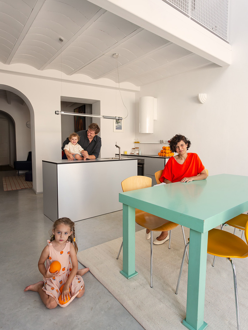 Family in modern kitchen with colorful table and chairs, child holding orange, warm and inviting home atmosphere.