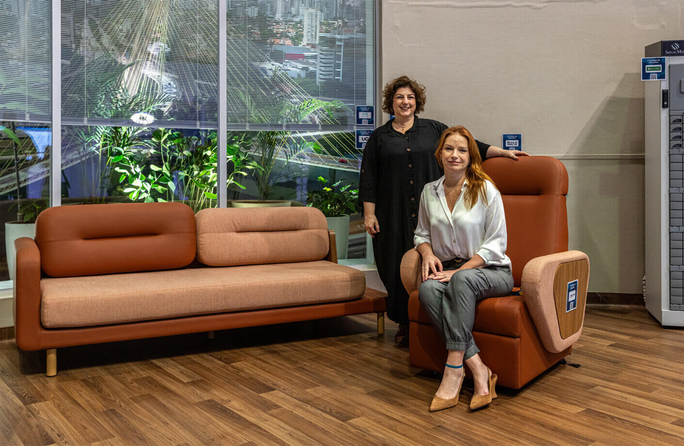 Two women showcase modern furniture in a stylish room with plants and city view.