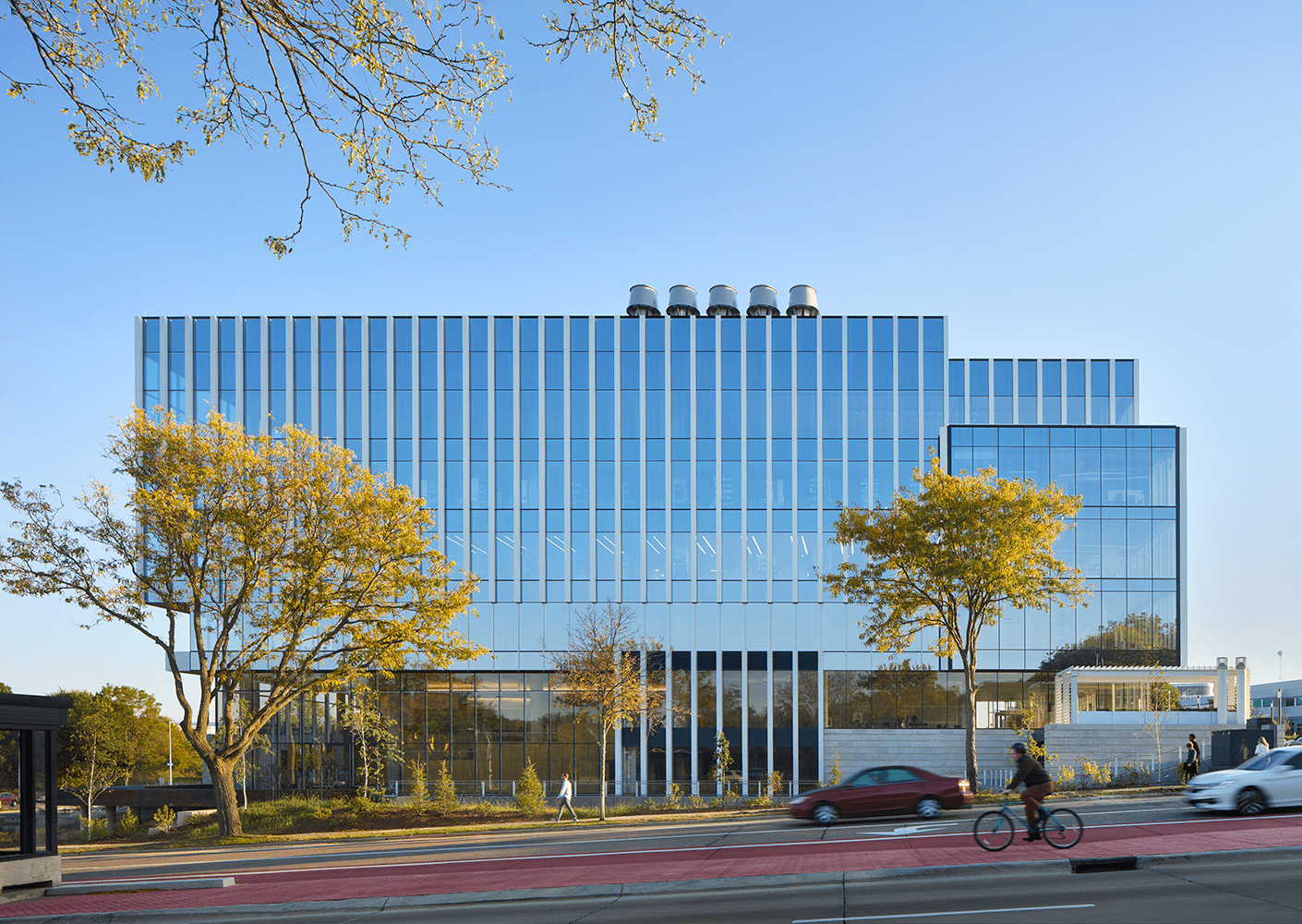 Modern glass building with trees, cars, and a cyclist on a sunny day.