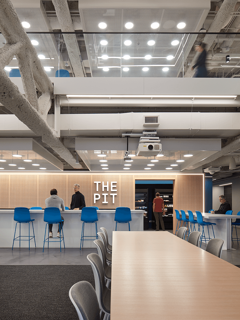 Modern open-plan office space with blue chairs and people at a counter labeled The Pit.