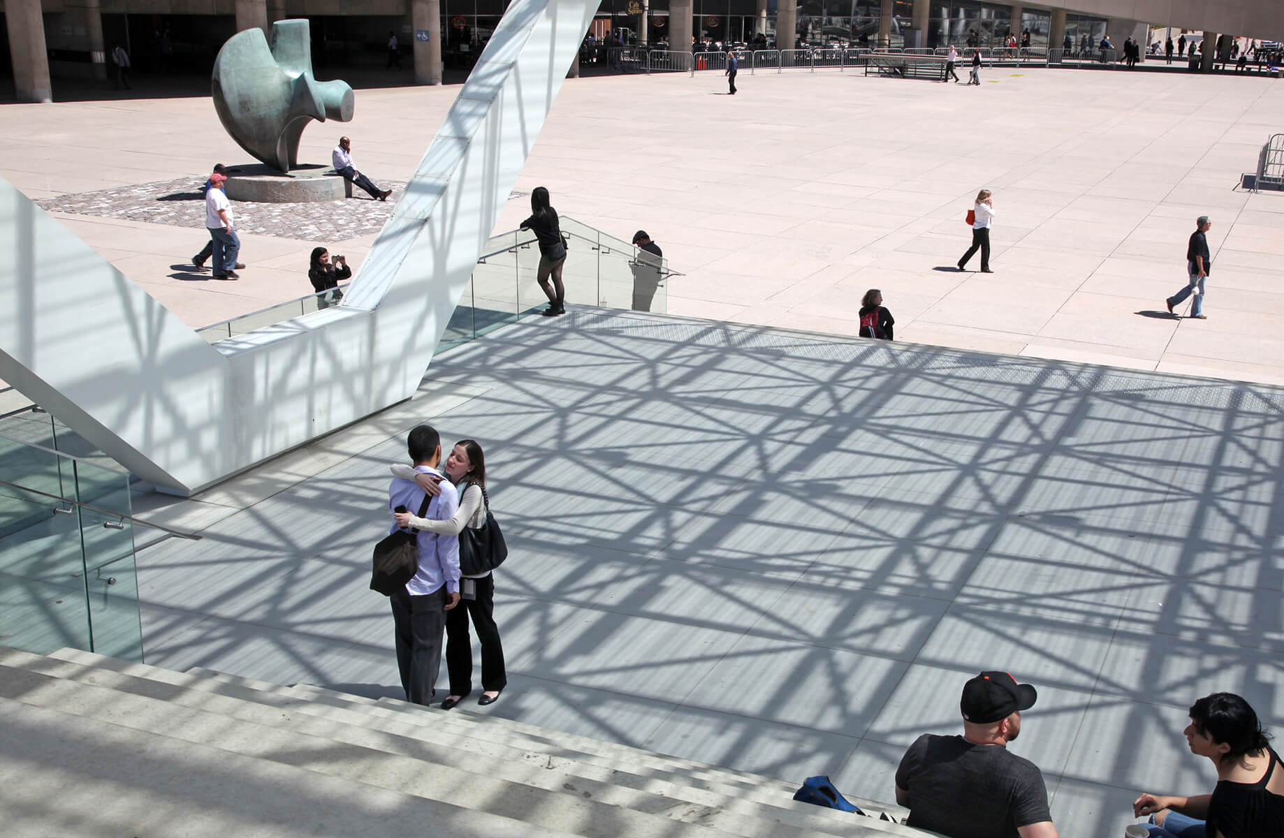 Modern plaza with art sculpture and people interacting under geometric shadows.
