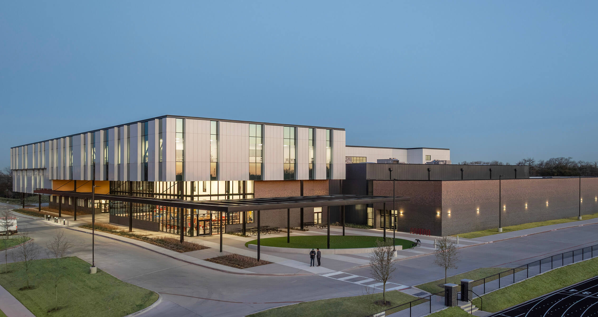 Modern school exterior at dusk with large windows and landscaped surroundings. Mix of white paneling on one side and darker brick on the other.