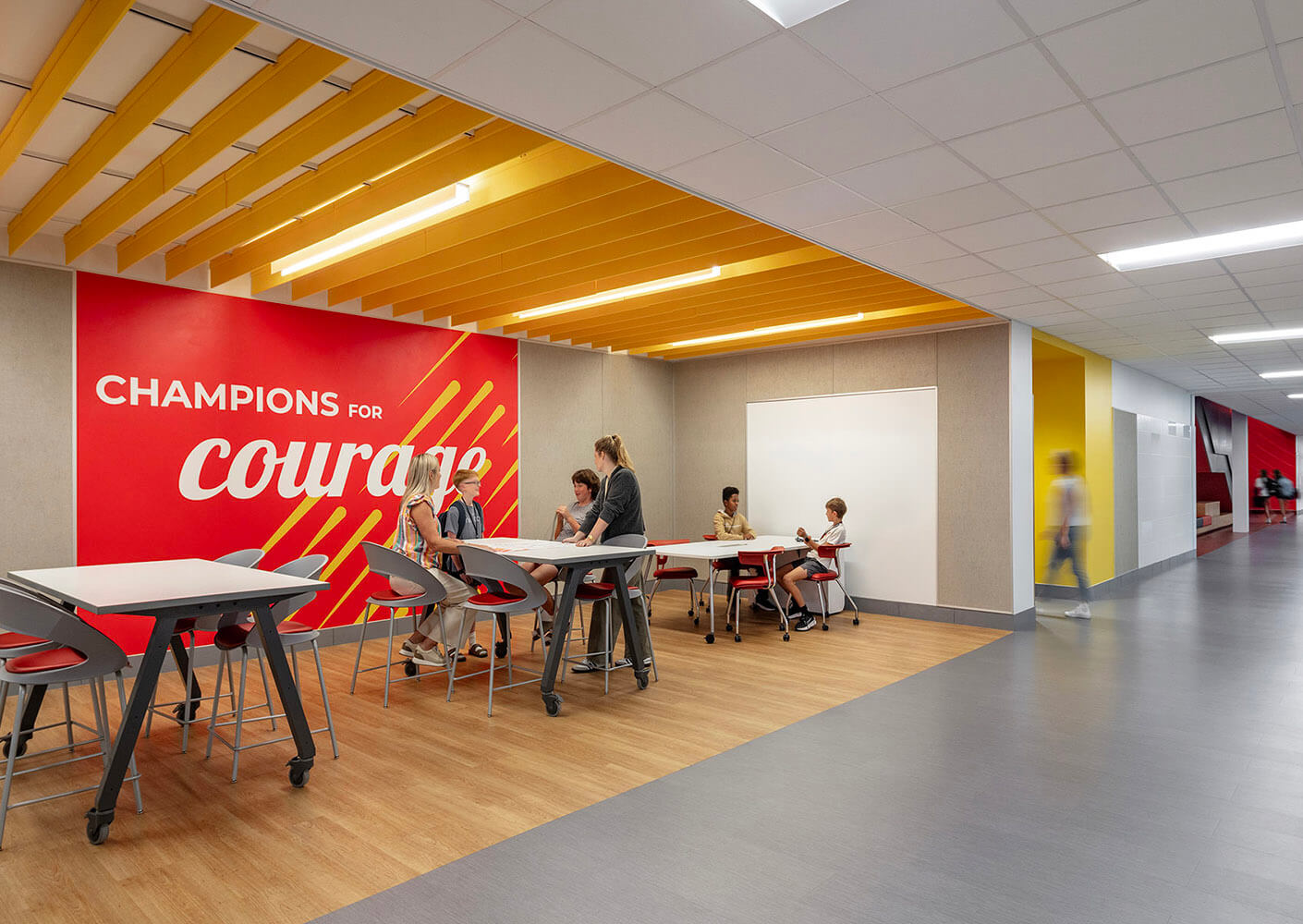Modern school lounge open to the hallway, with students at tables under a yellow ceiling and by colorful graphics including the phrase 