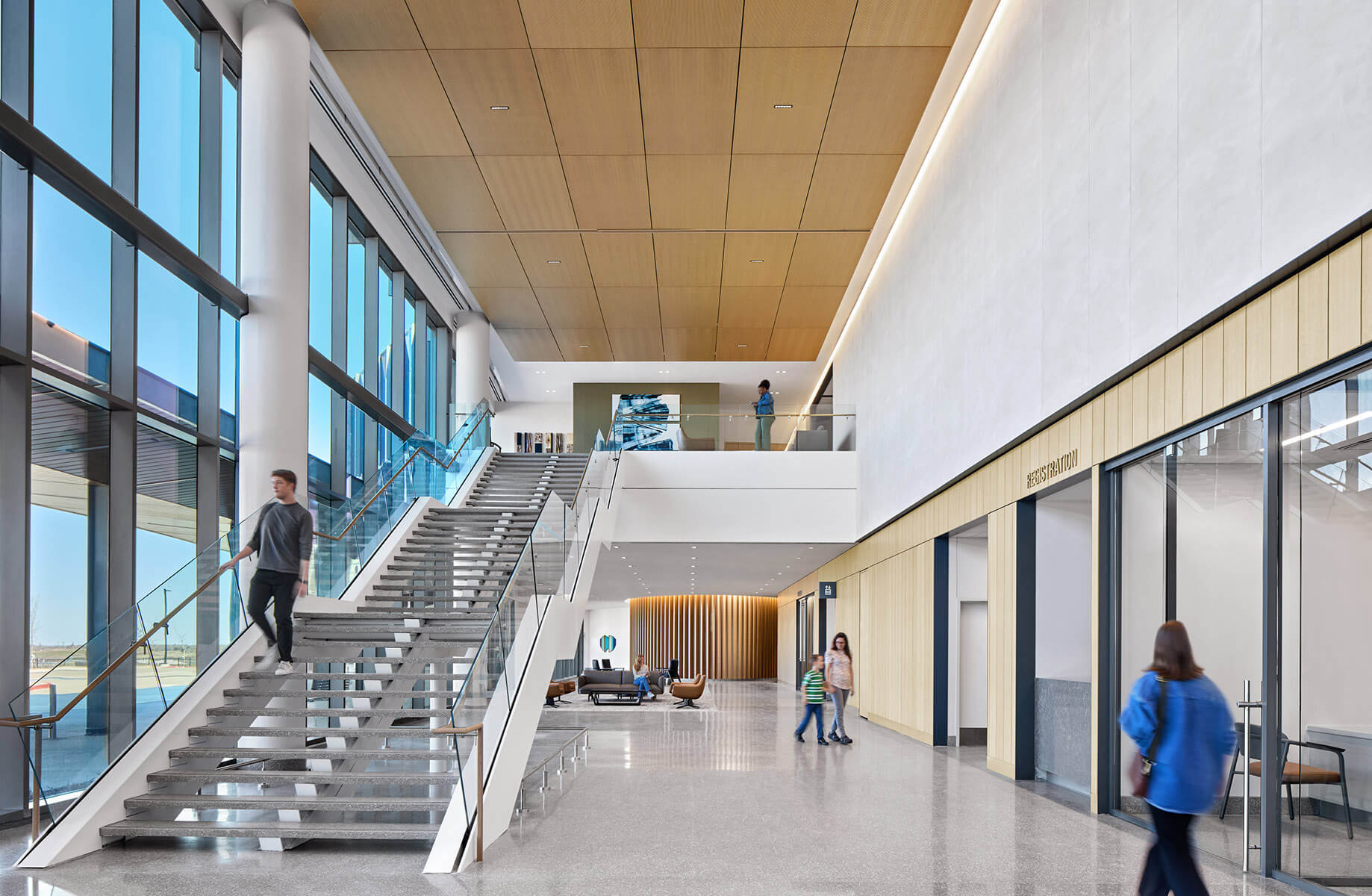 Modern lobby with tall windows, a grand staircase, and people walking through.