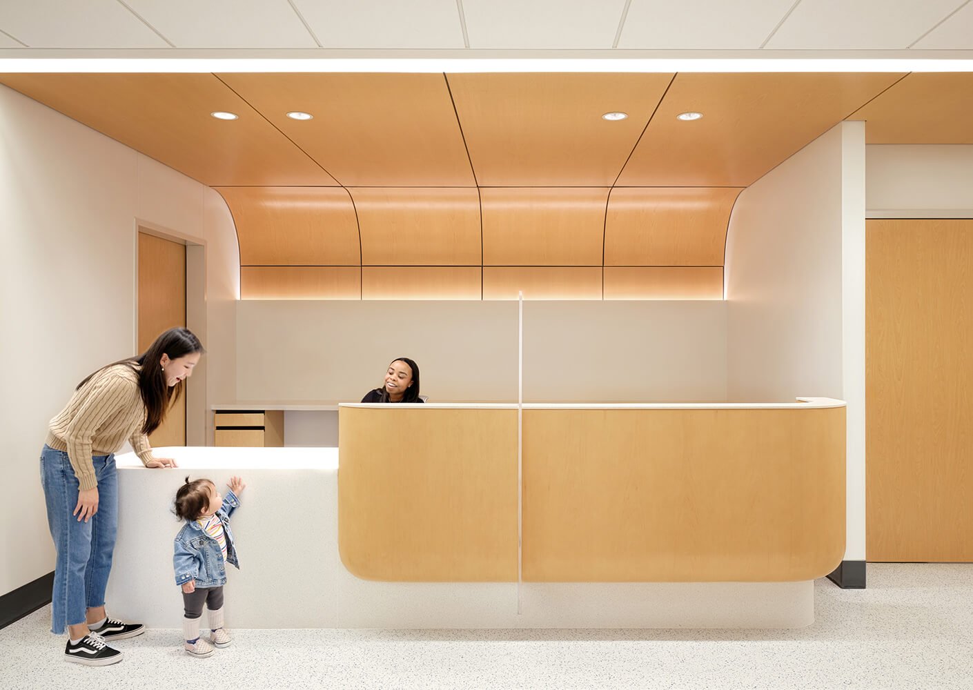 Modern reception area grounded in warm wood accents, featuring woman, child, and smiling receptionist.