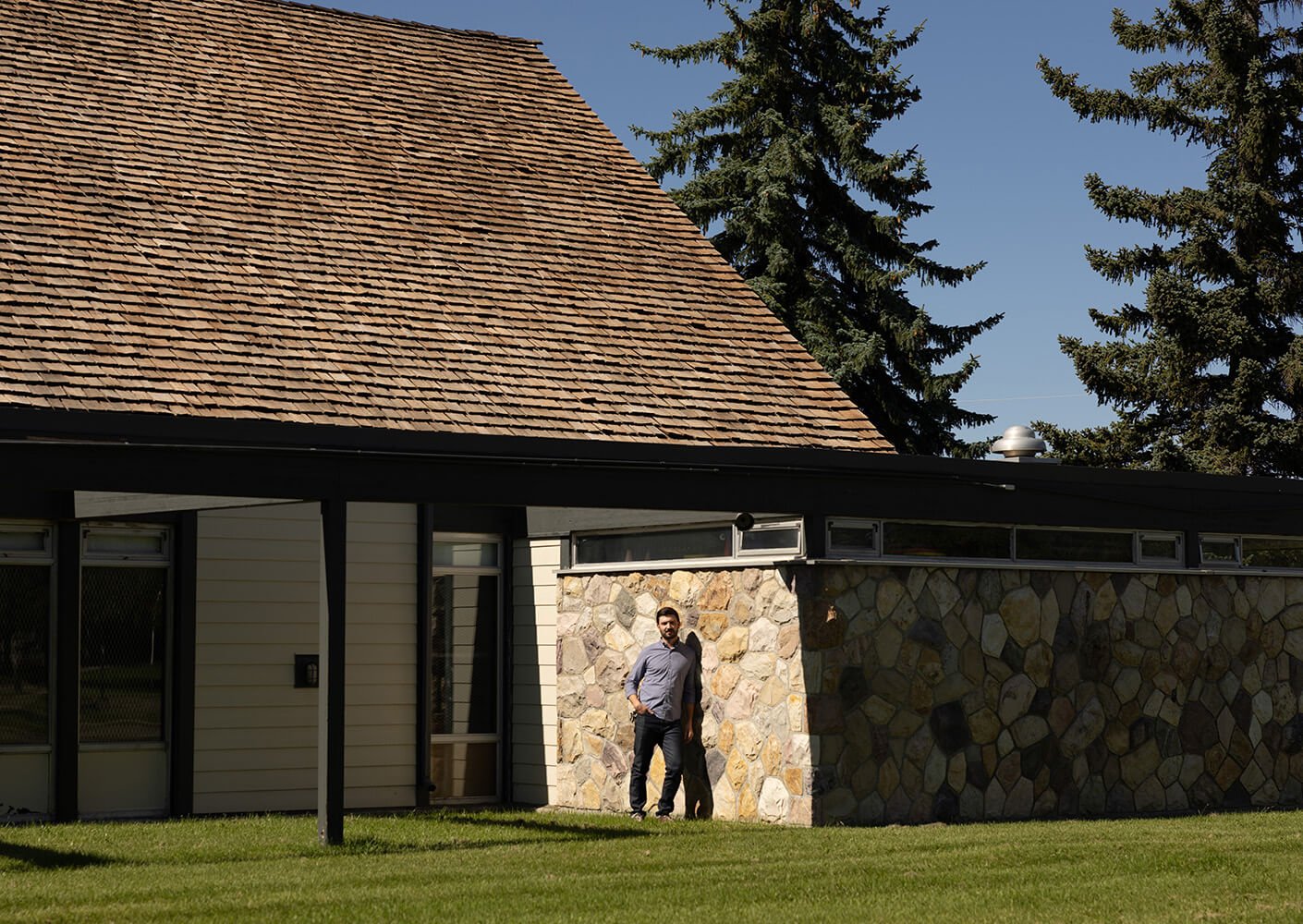 A man standing outside a stone and wood building with a large shingle roof.