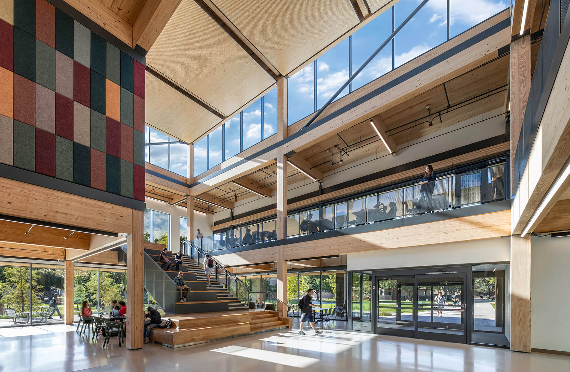 Modern open atrium with wooden beams and large windows, people studying and walking.
