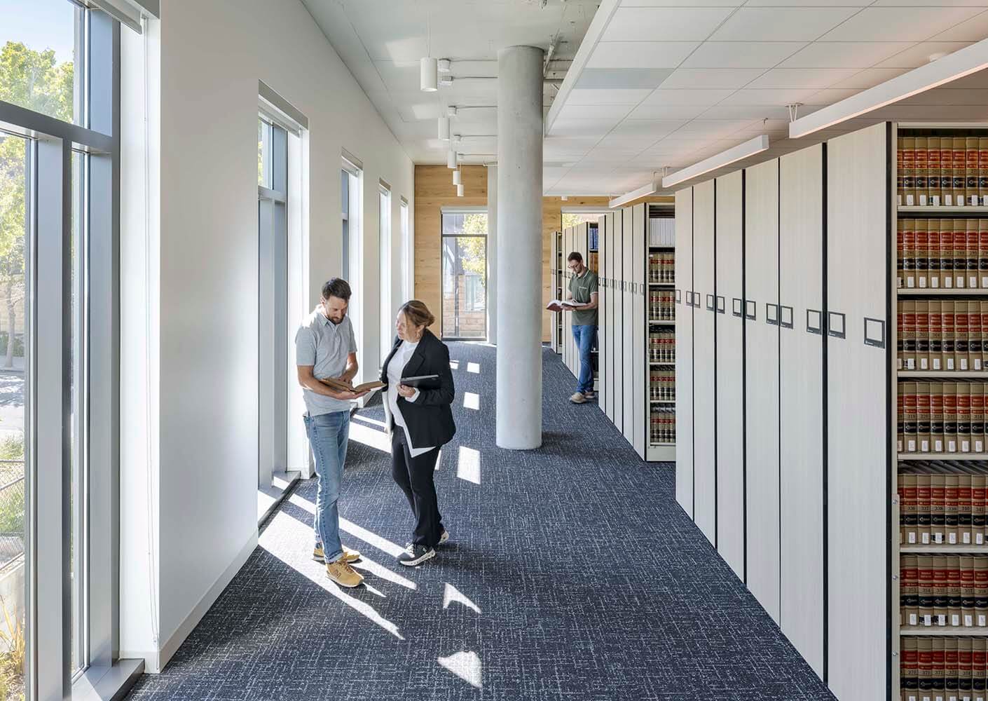 People discussing next to bookshelves in a sunlit modern office hallway.