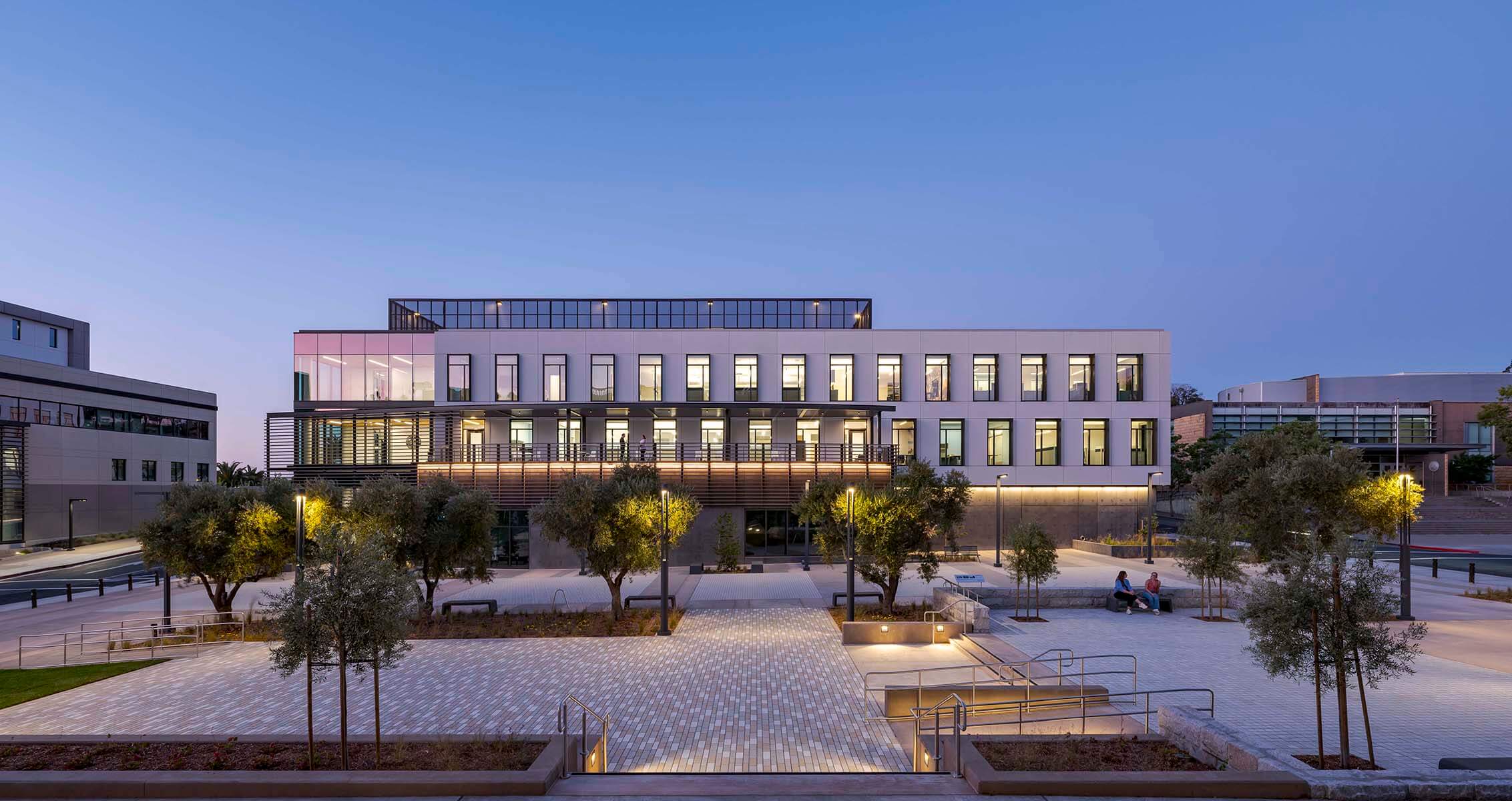 Modern building exterior at dusk with illuminated windows and surrounding trees.