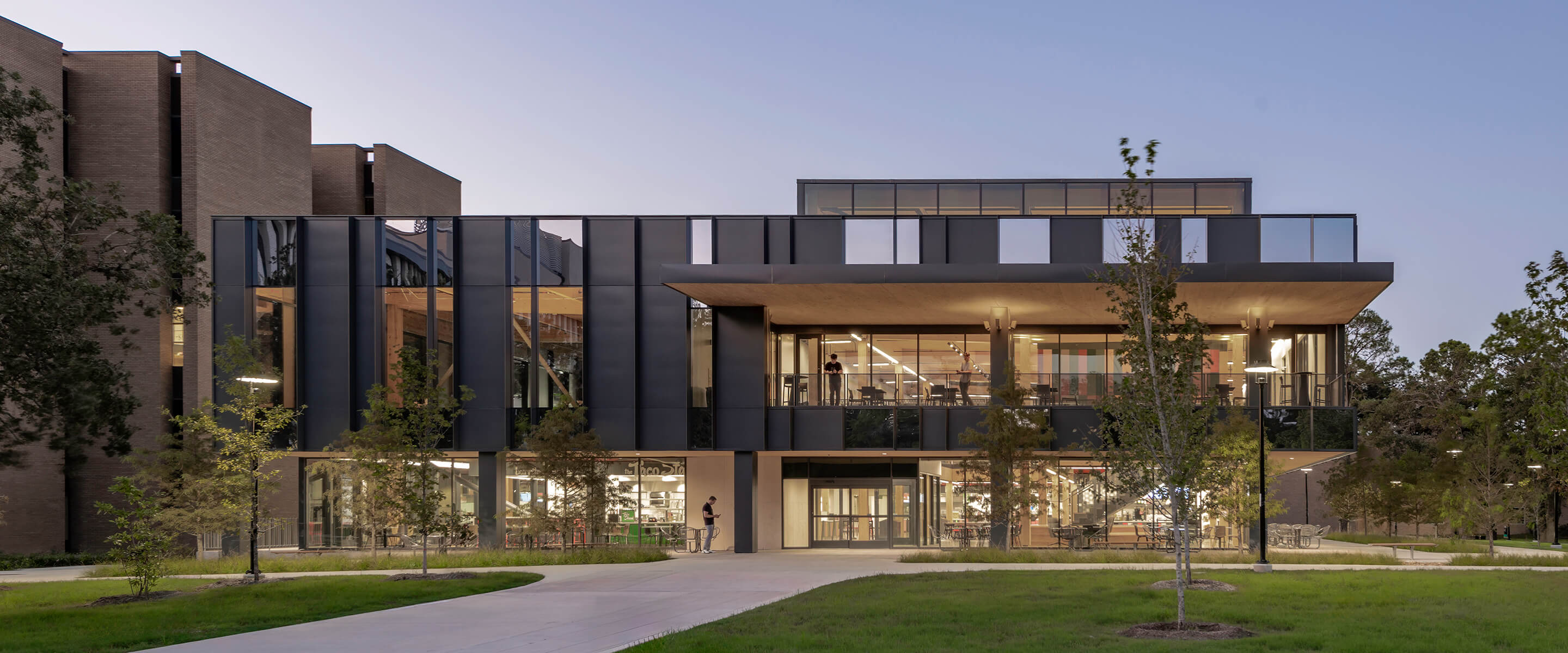 Modern building exterior at dusk with large windows and surrounding greenery.