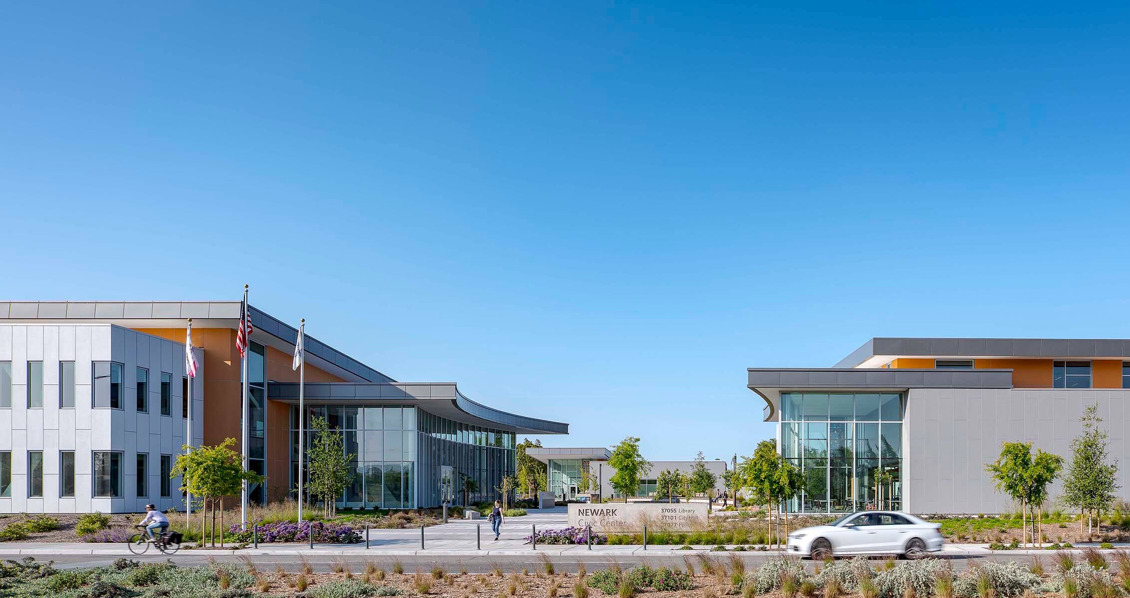 Modern civic center building with flags, cyclist, and car under clear sky.