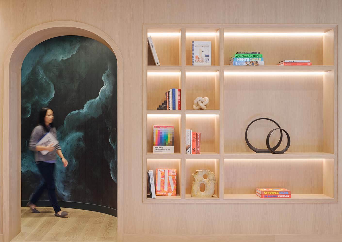 Modern interior with a woman walking by an illuminated wooden bookshelf.