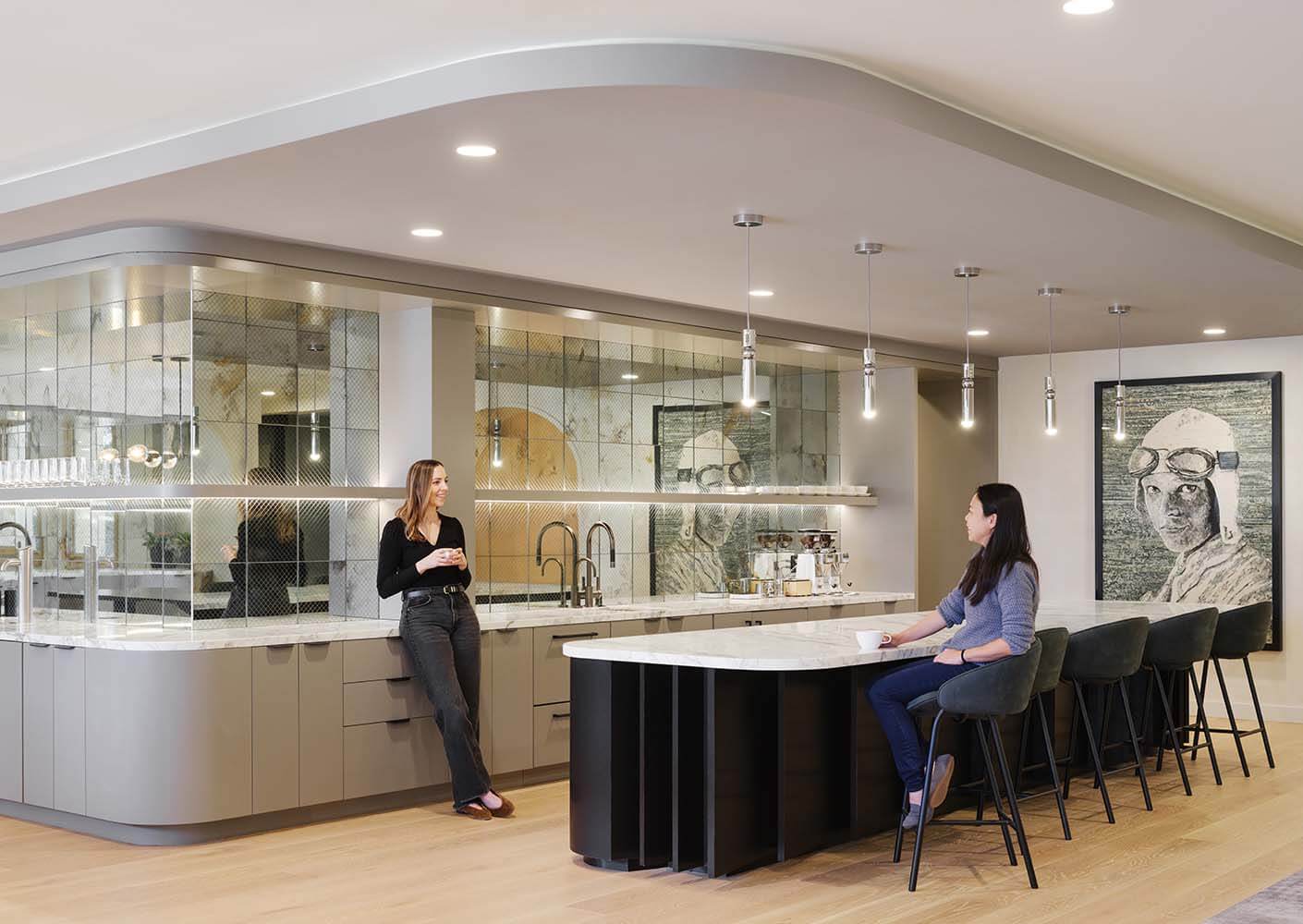 Modern office kitchen with two women talking, stylish design, wall art, bar seating.