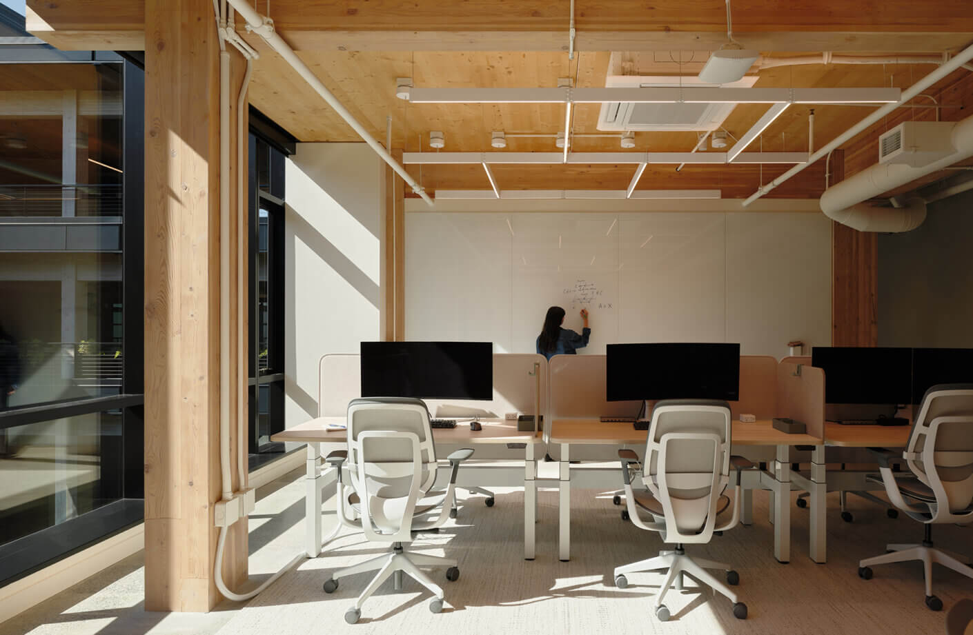 Modern office space with desks, chairs, and person writing on whiteboard.