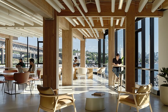 Modern coworking space with natural light, wooden beams, and people working by large windows.