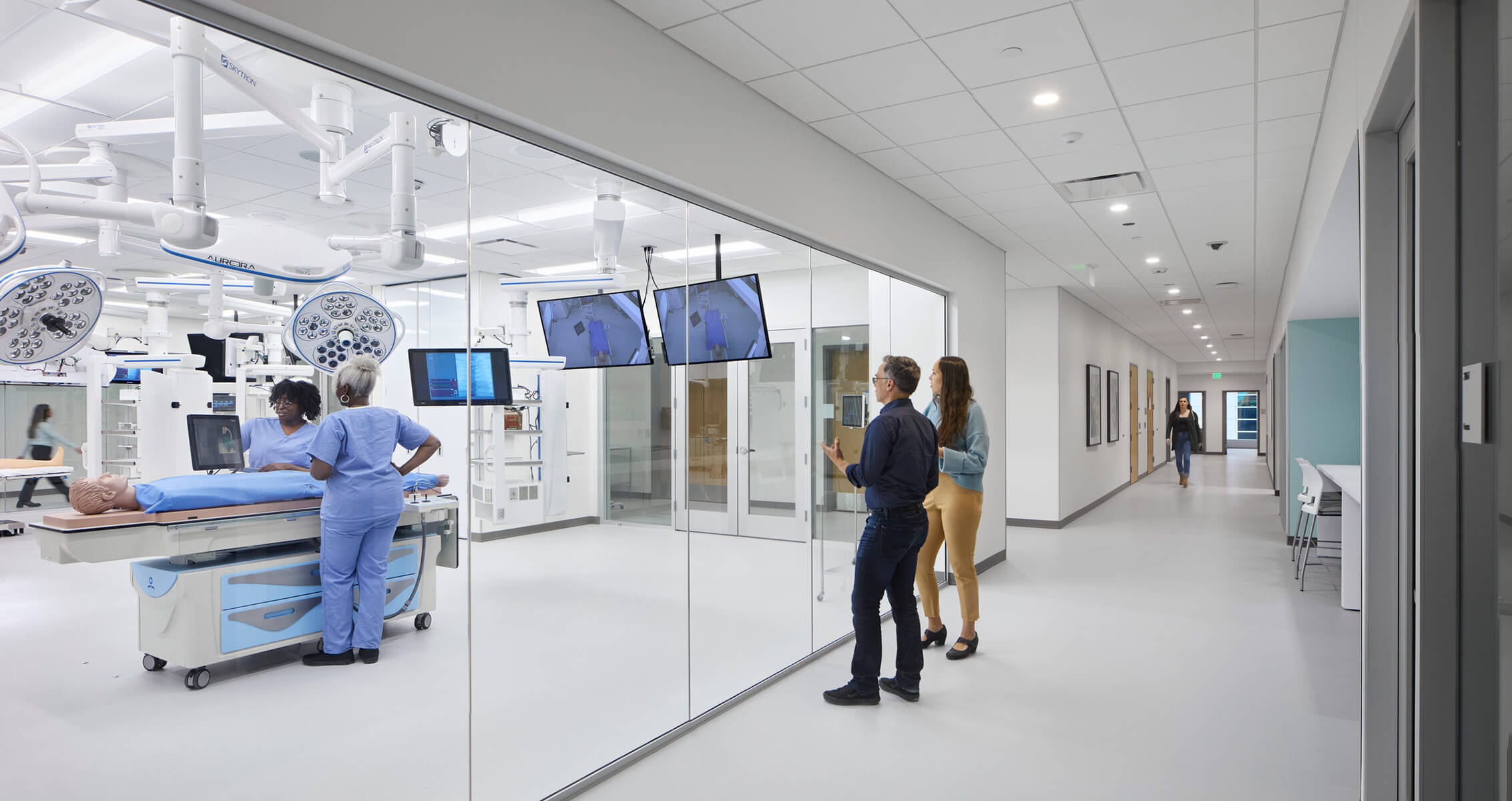 Medical professionals in scrubs with a patient in a hospital examining room with imaging screens and a glass wall