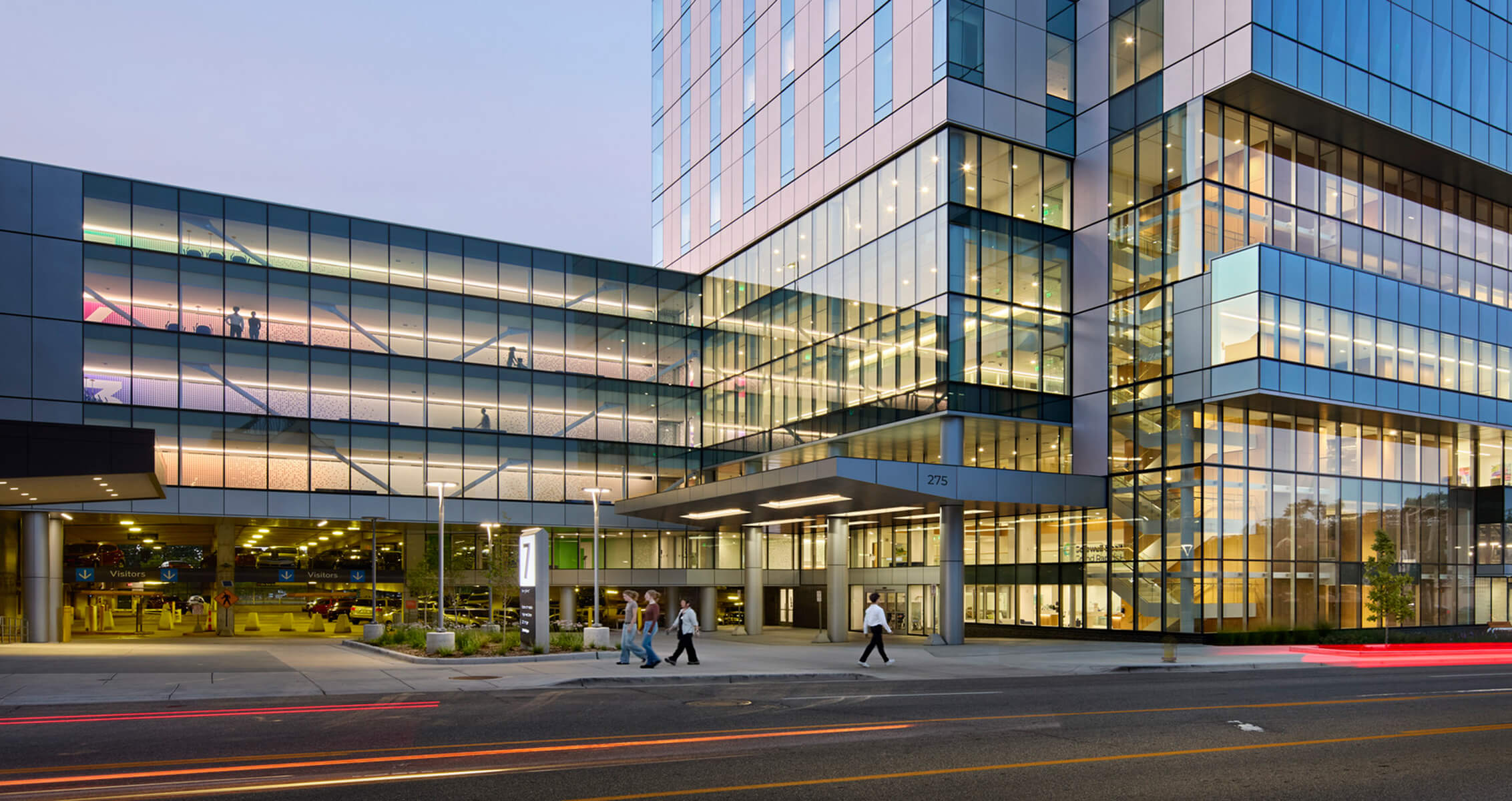 Exterior of a hospital with large glass full-length windows lit up in the evening; people walk outside the entrance and look down from the windows above