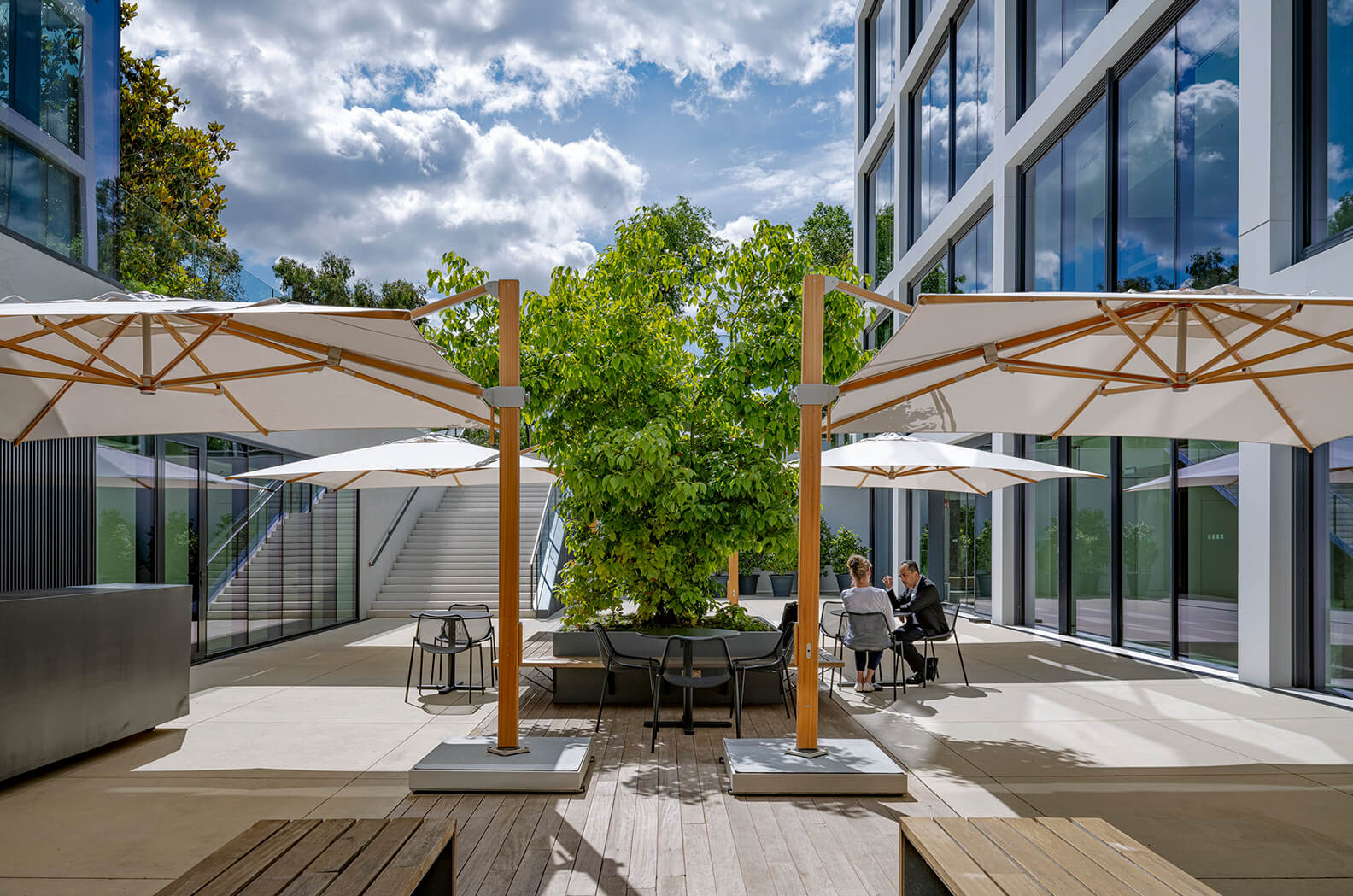 Modern office terrace with greenery and large umbrellas under a partly cloudy sky.
