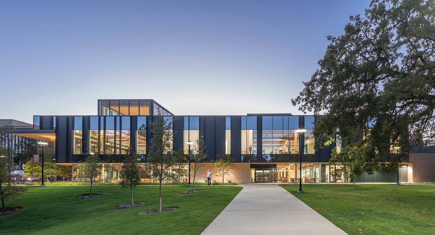 Modern campus building with glass facade at dusk, surrounded by green lawns and trees.