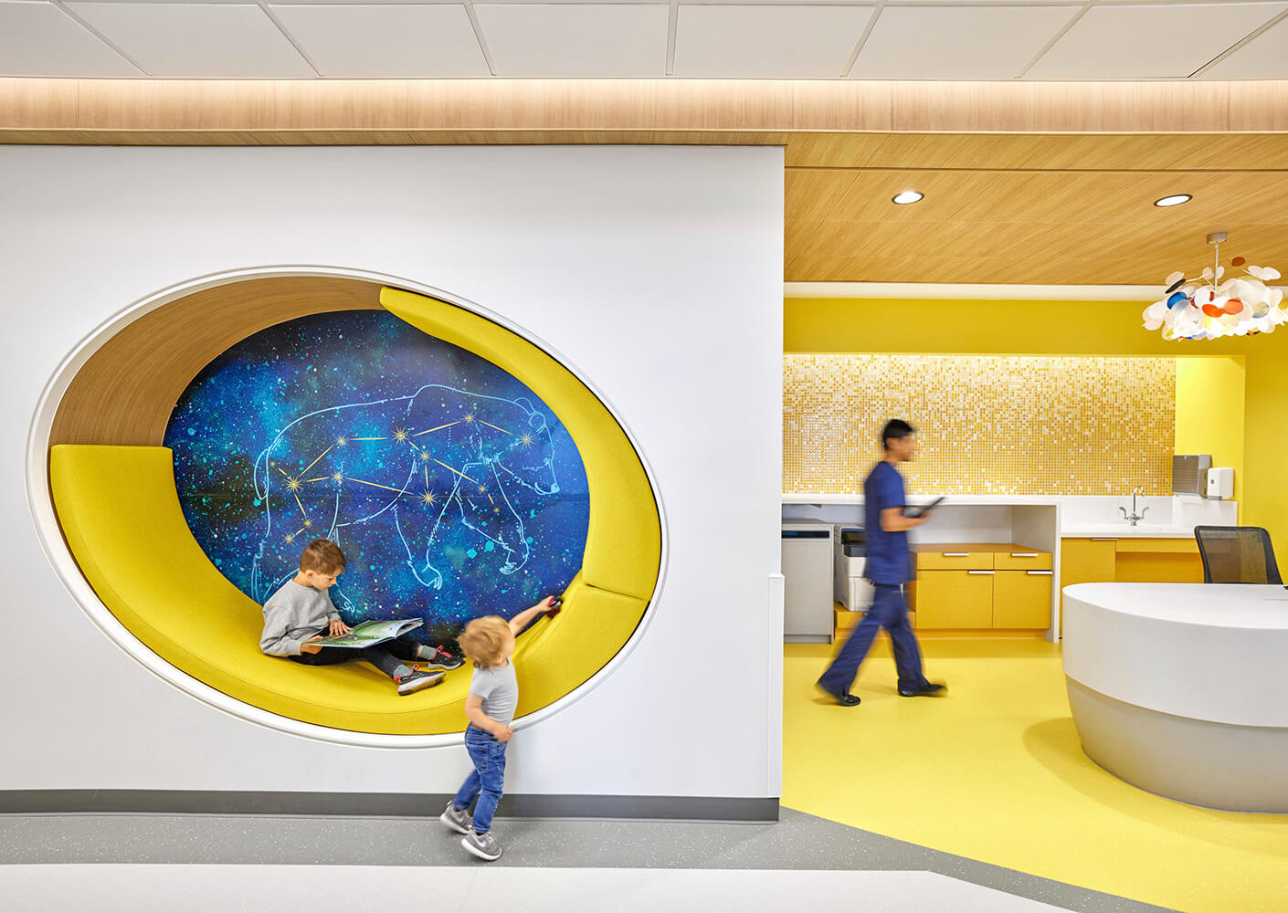 Children's reading space inset in wall with constellation mural. A person walks in the background in bright yellow area of medical clinic.