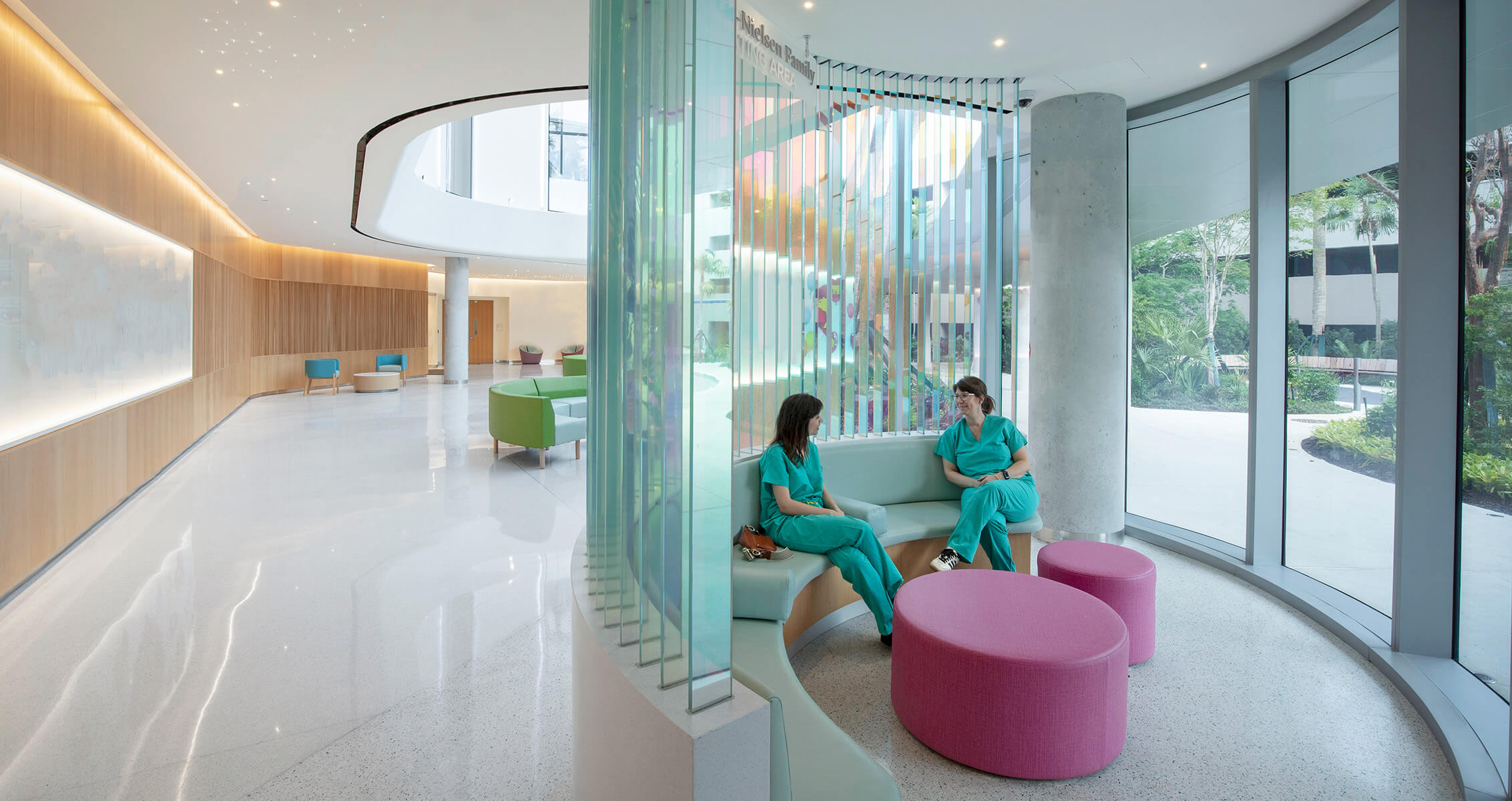 Two healthcare workers in scrubs converse on comfortable seating in a hospital lounge interior with curved walls and colorful furniture