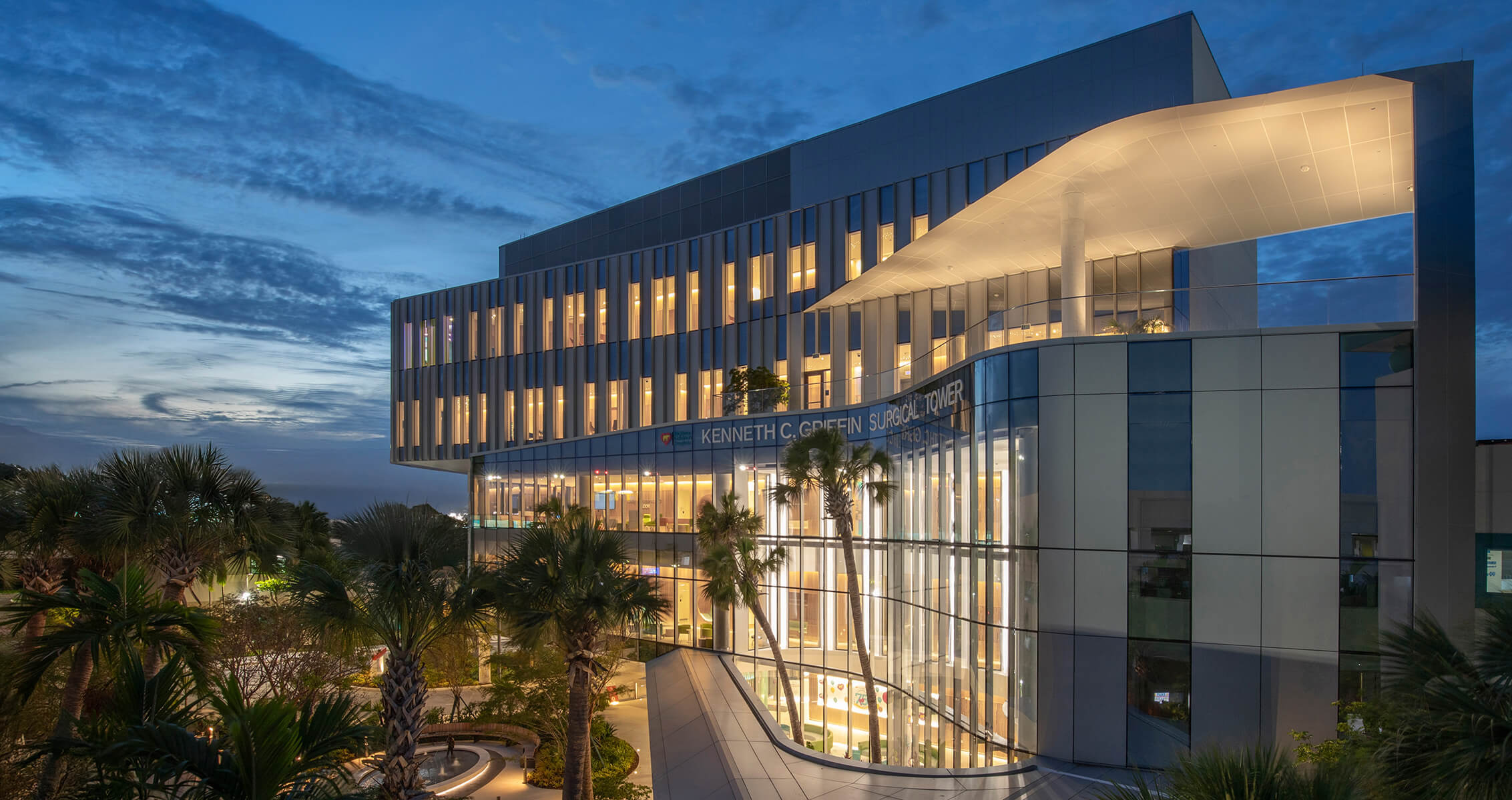 Exterior of a hospital at night with many lit windows and palm trees in the foreground