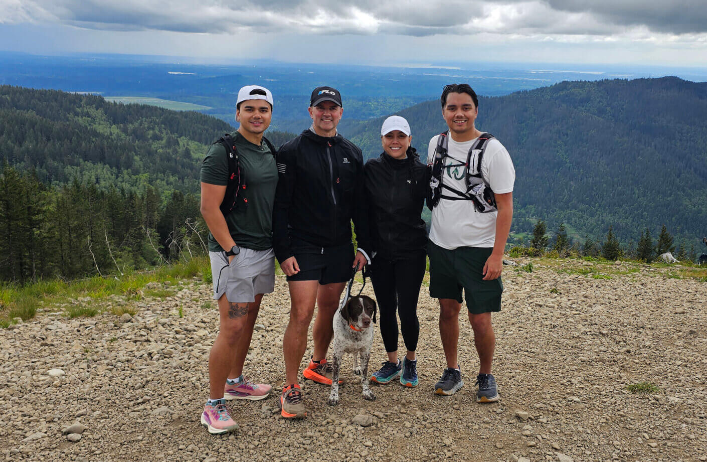 Group hiking with dog on scenic mountain trail with lush forest and cloudy sky.