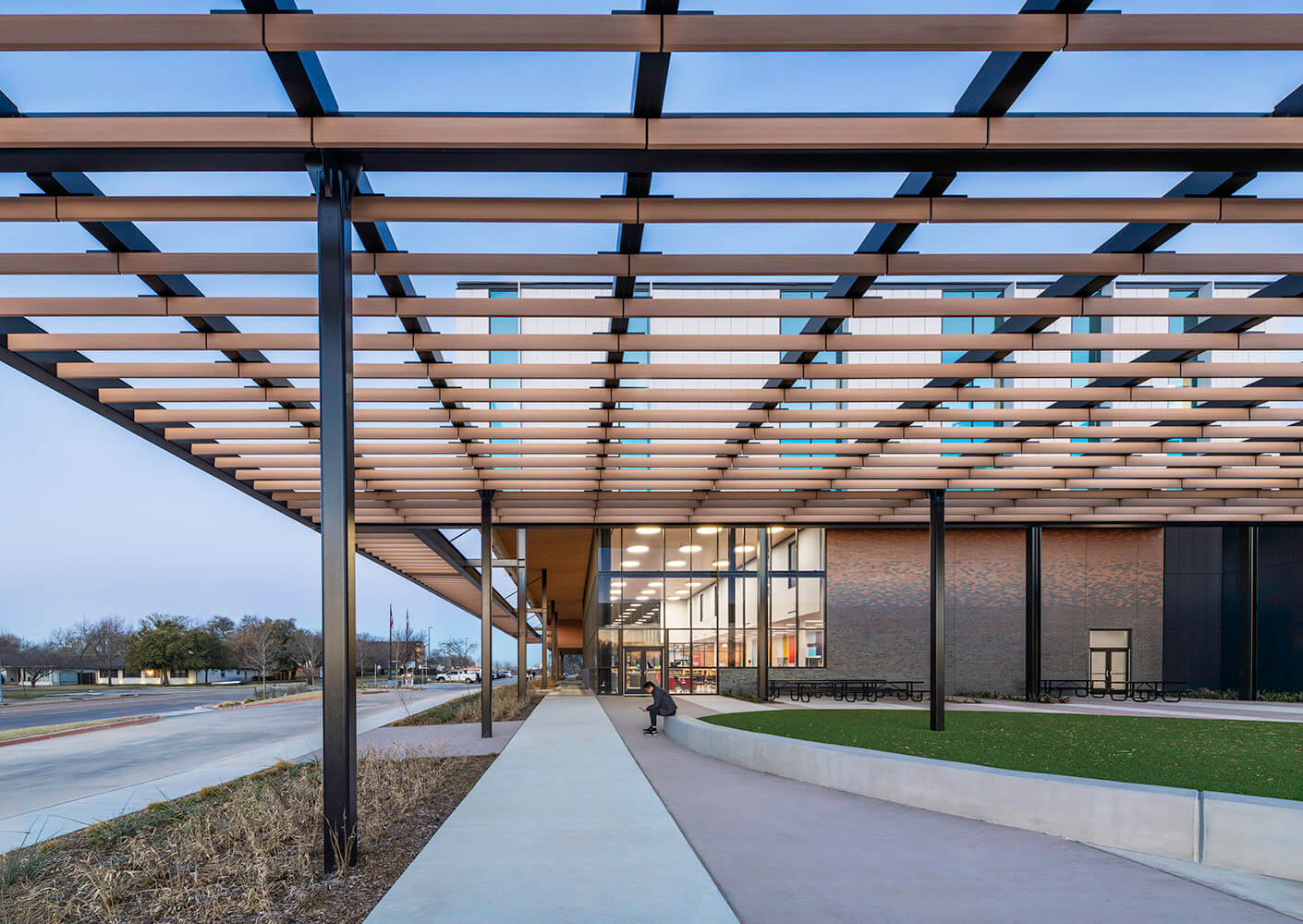 Modern building with wooden beam pergola and glass corner facade at dusk. Brick transitions from darker at the base to lighter higher up.