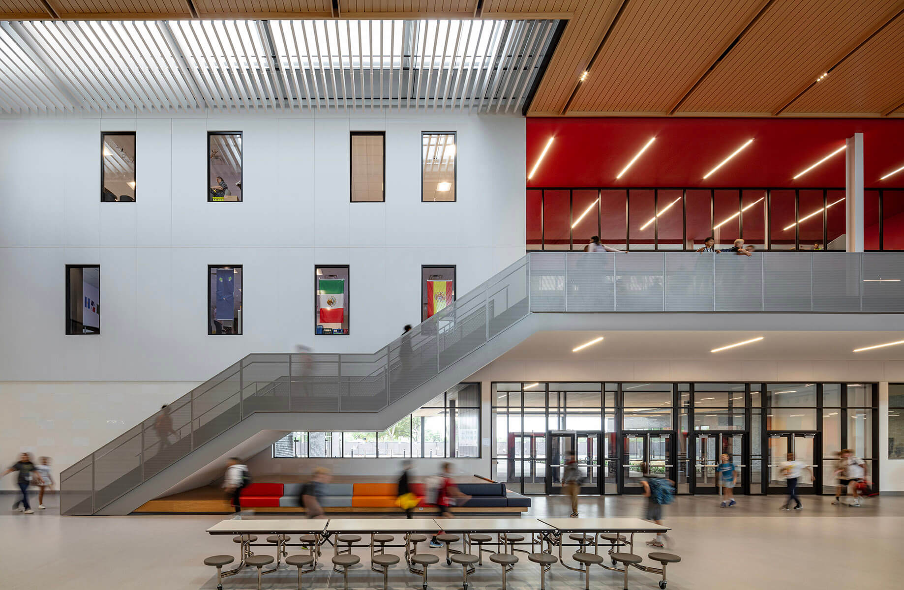 Modern school interior with flags, stairs, and students walking in a bright common area under a light well.