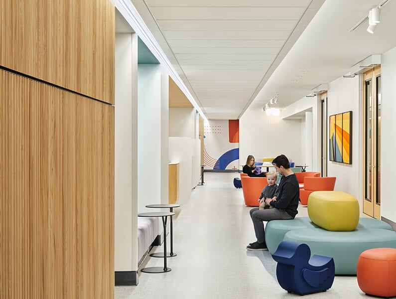 Brightly lit modern lounge area with colorful seating, parent relaxing with child in lap, and geometric wall art.
