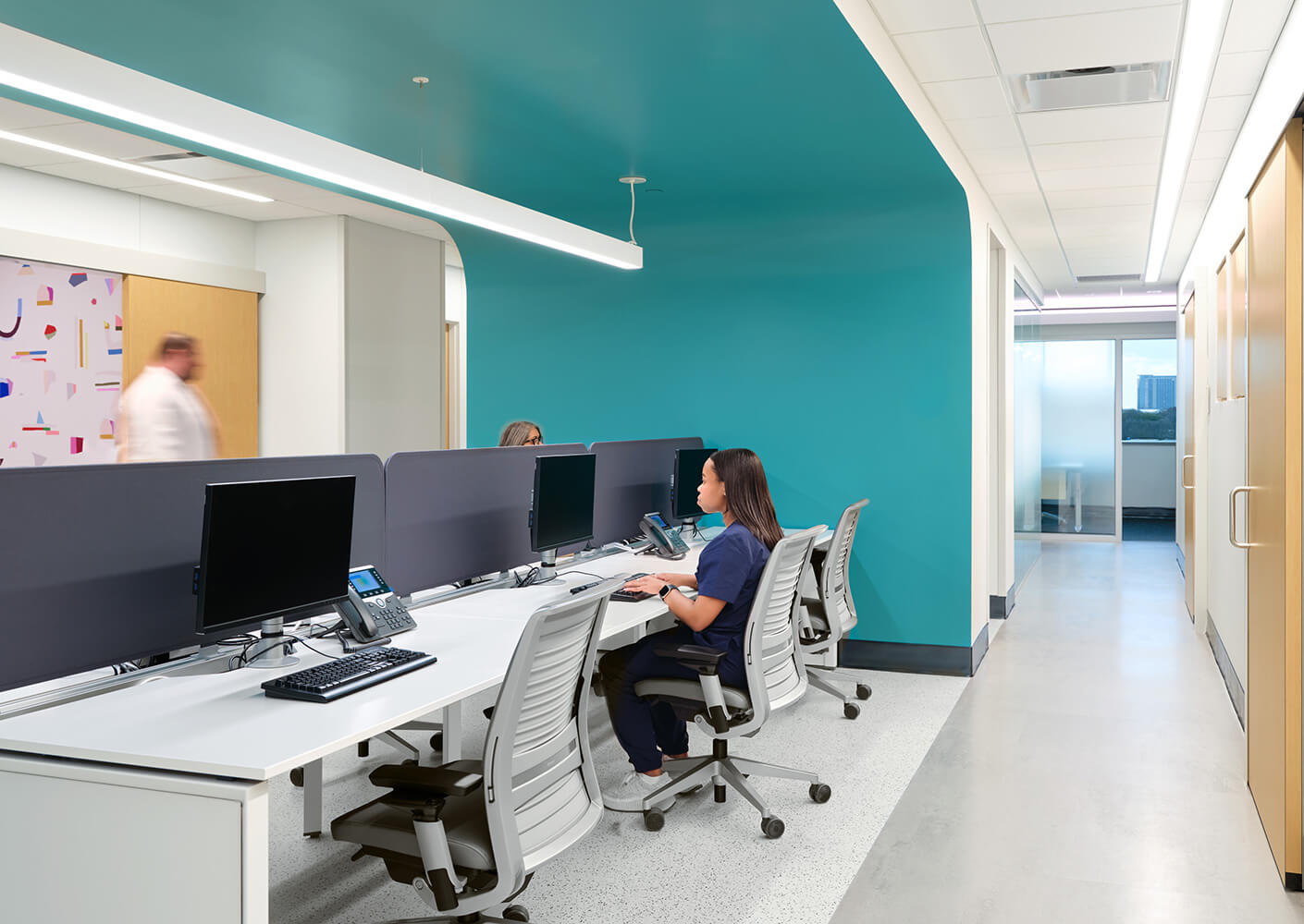Physician workspace with teal walls, desks and computer, open to two hallways lined with doors; person in scrubs working.