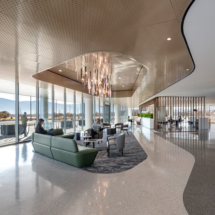 Lobby with seating under a chandelier and people walking along a curved path next to an undulating wood wall and floor-to-ceiling windows.