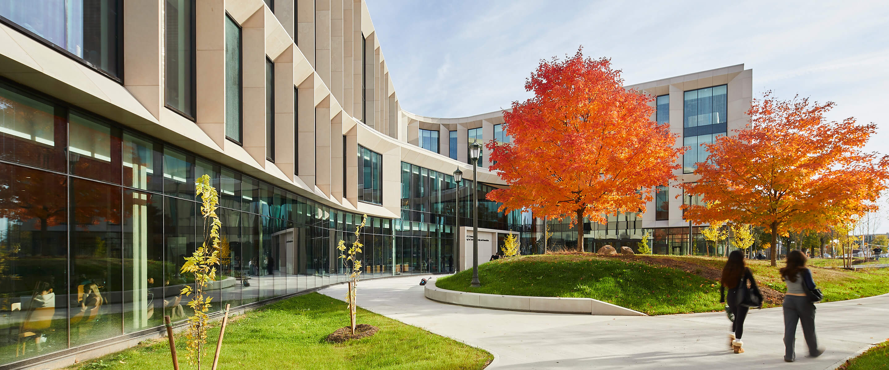 Modern building with autumn trees and two people walking nearby.