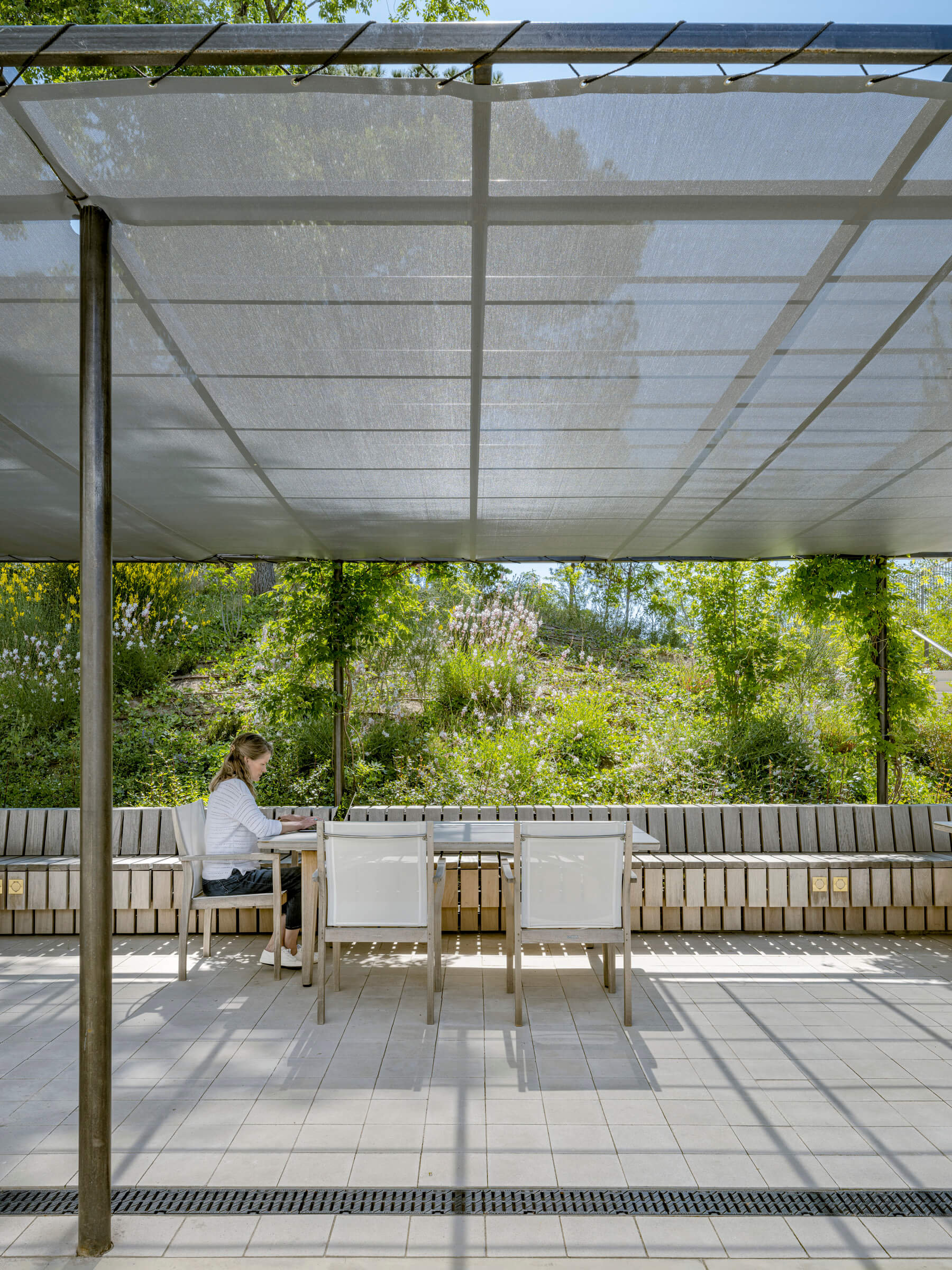 Person working at outdoor table under sunshade in lush garden.