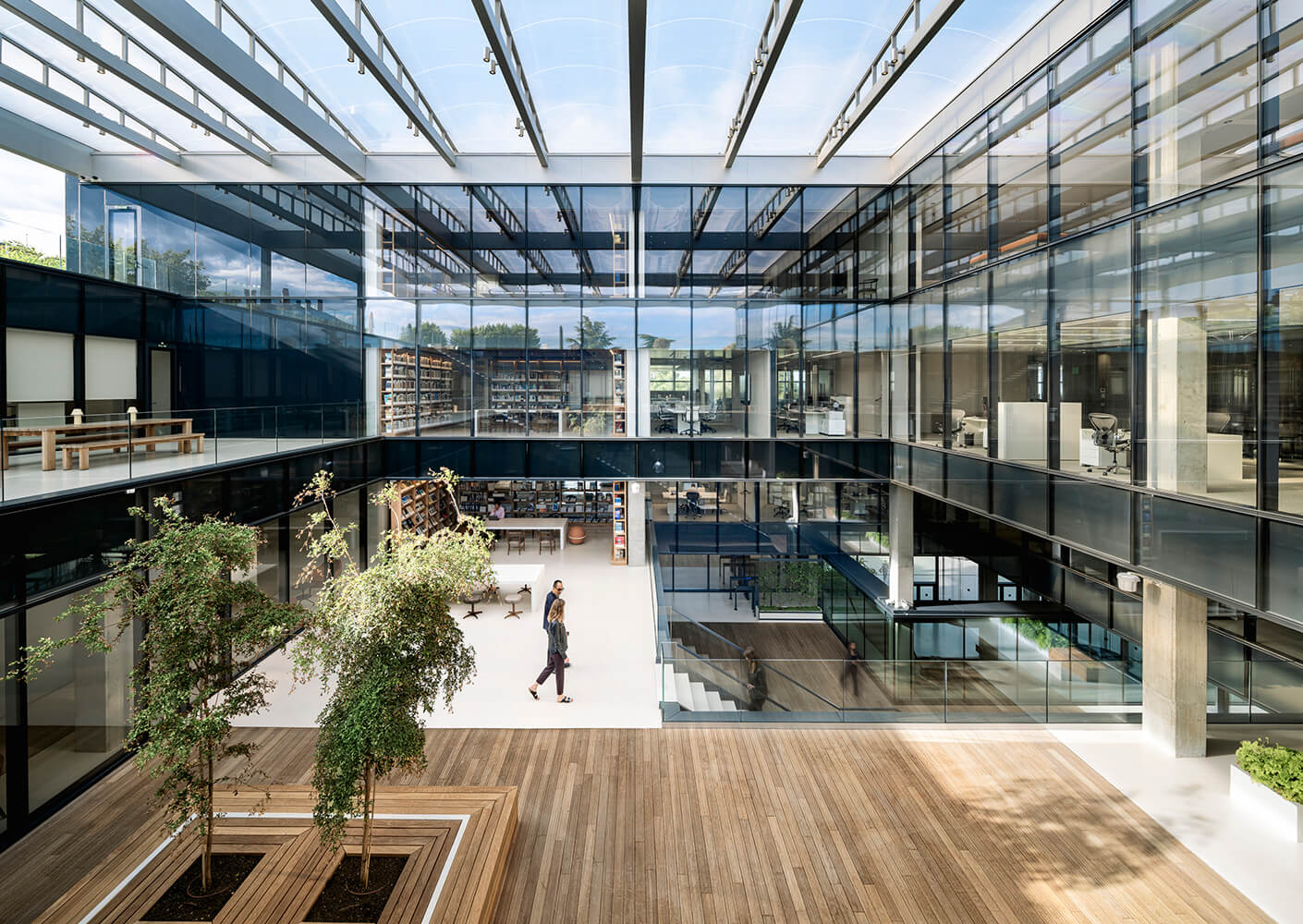 Modern office atrium with glass walls, greenery, and a person walking.
