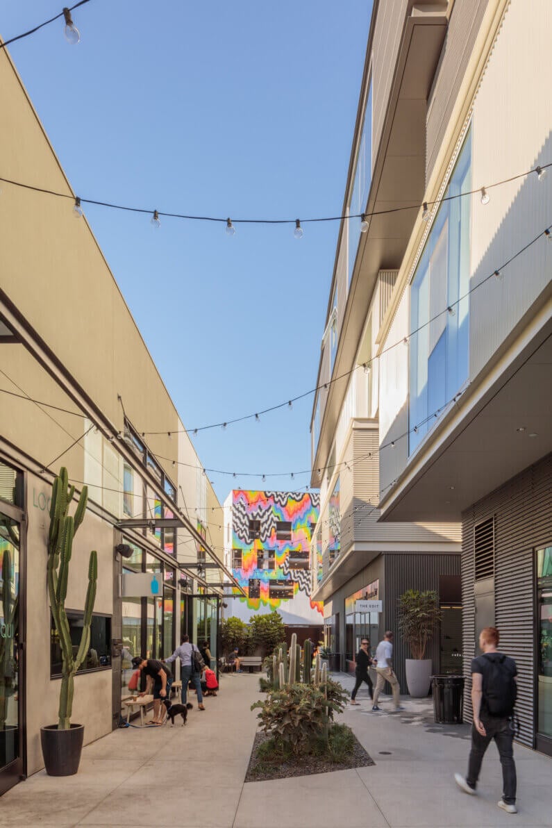 Colorful urban alley with string lights, mural, people, and cactus plants under clear sky.