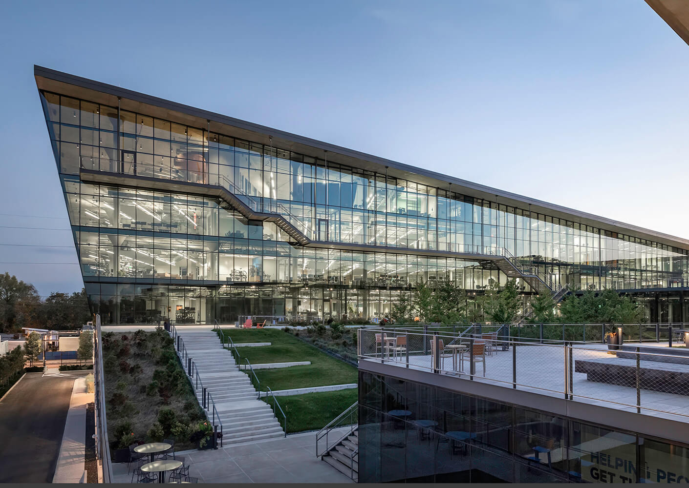 Modern glass office building with stairs and greenery at dusk.