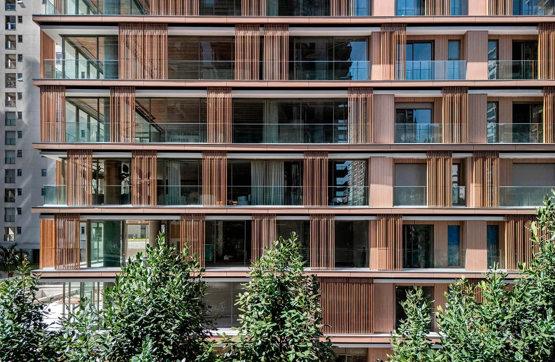 Modern building facade with glass balconies and wooden slats, surrounded by greenery.