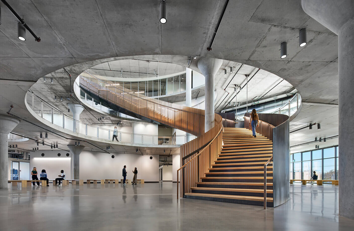 Modern architectural interior with curved wooden staircase and people walking and sitting.