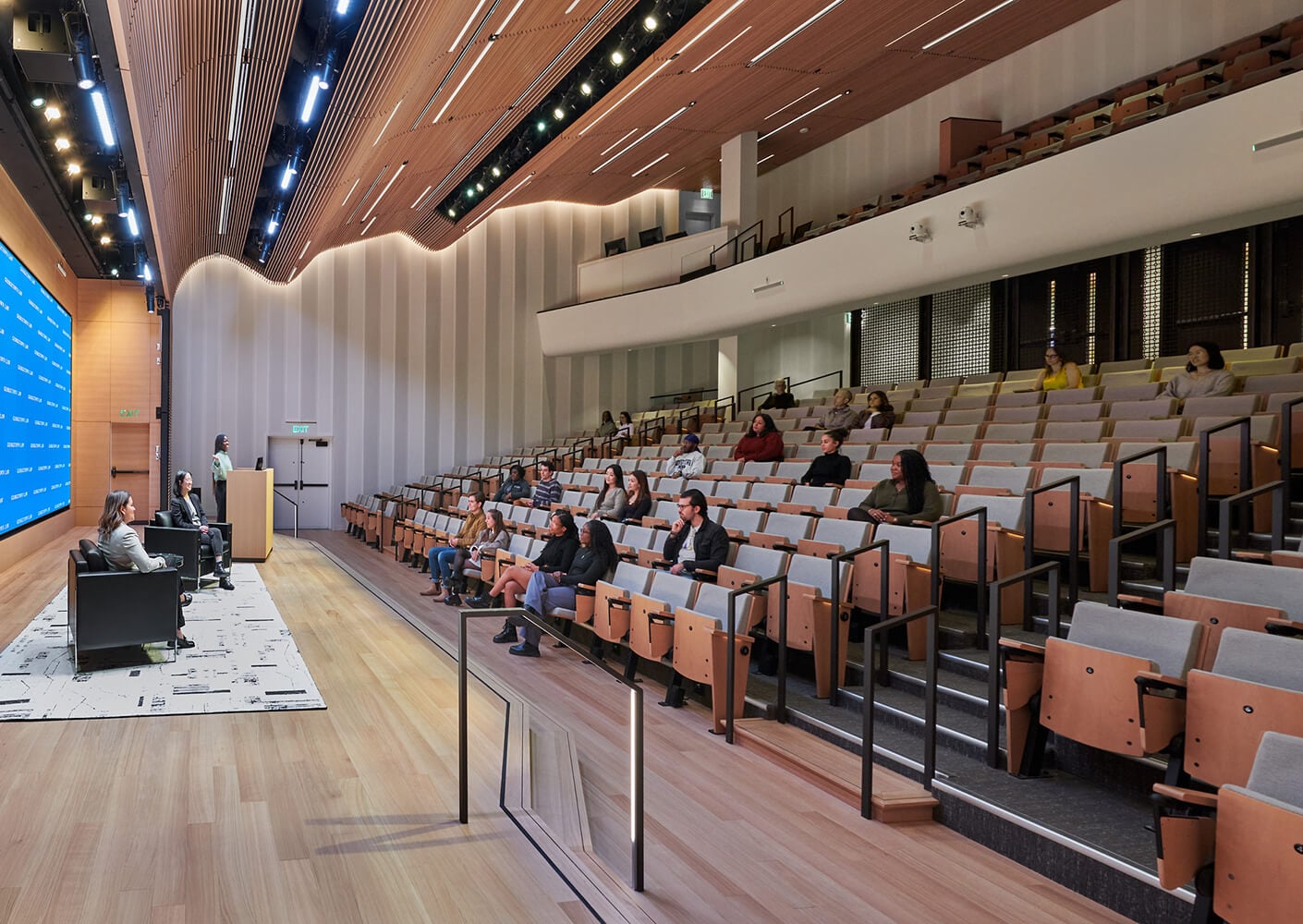 Modern auditorium with a panel discussion and sparsely filled seats.
