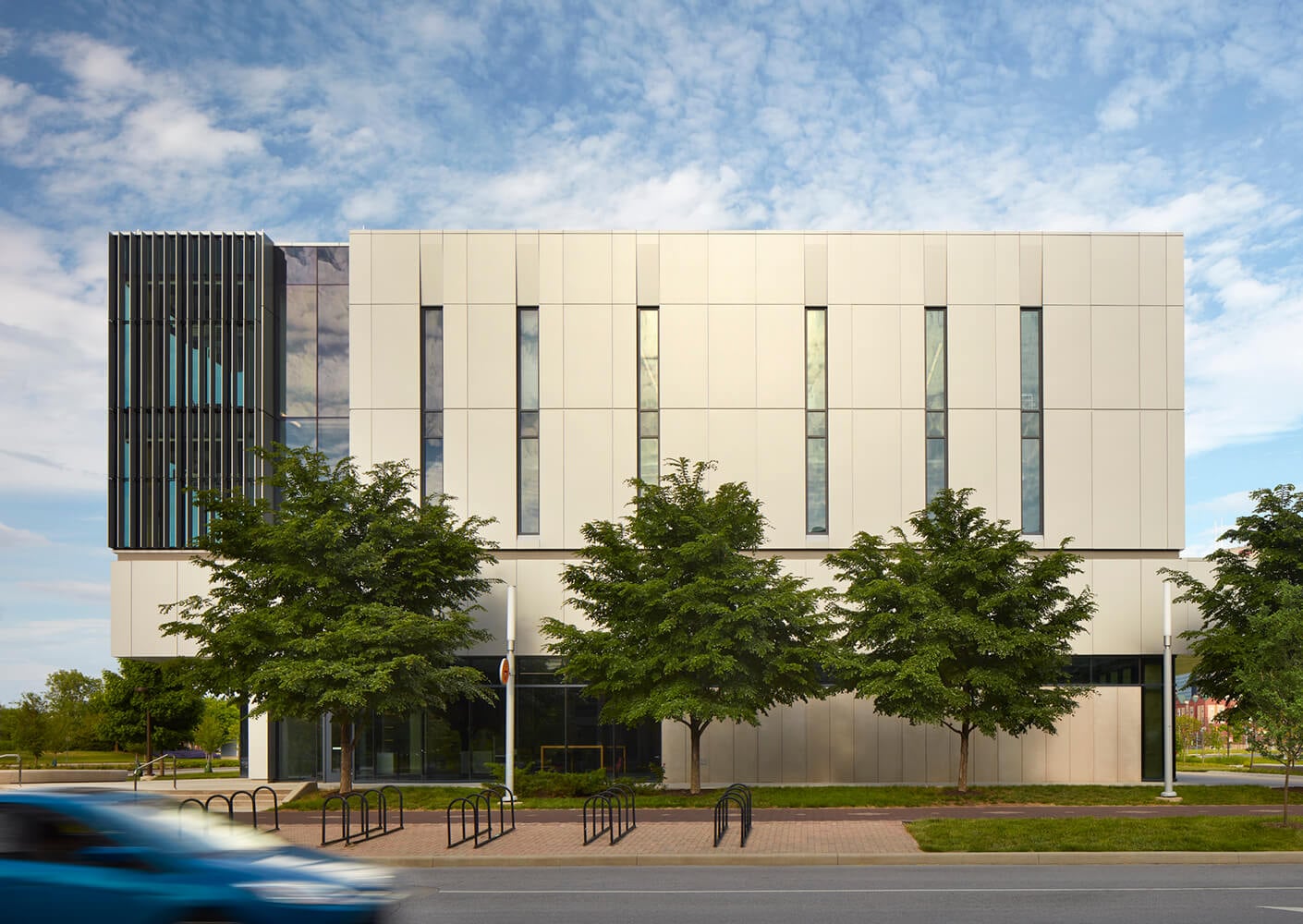 Modern white building facade with trees and a blurred blue car passing by.