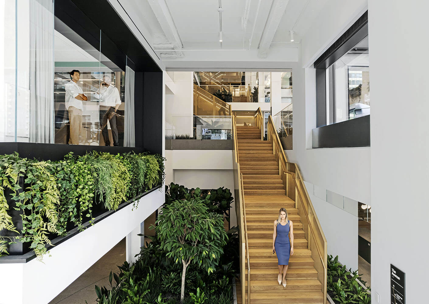 Modern office interior with lush greenery and a woman walking down wooden stairs.