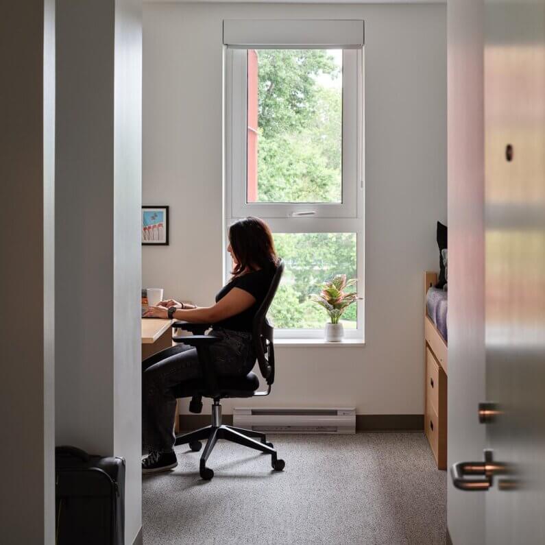 Person working at desk in a cozy bedroom with a window view.