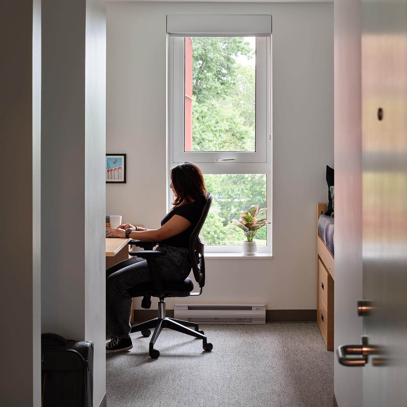 Person working at desk in a cozy bedroom with a window view.