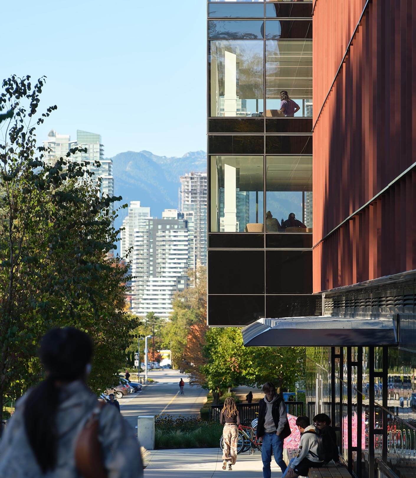 Modern urban street with people, glass building, and mountain views.