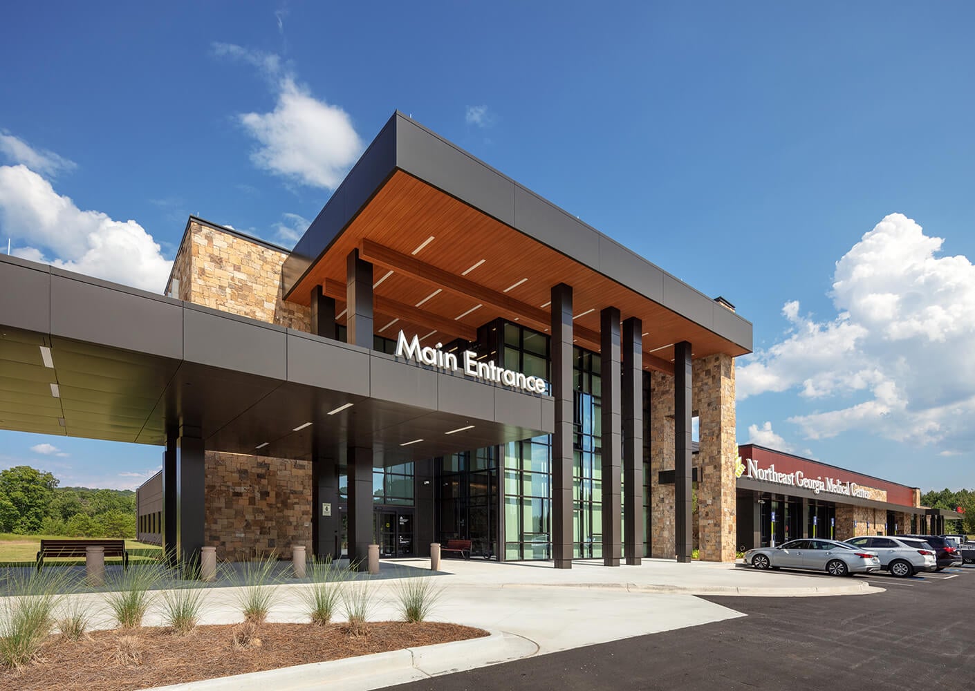 Modern hospital main entrance with cars parked outside under a clear blue sky.