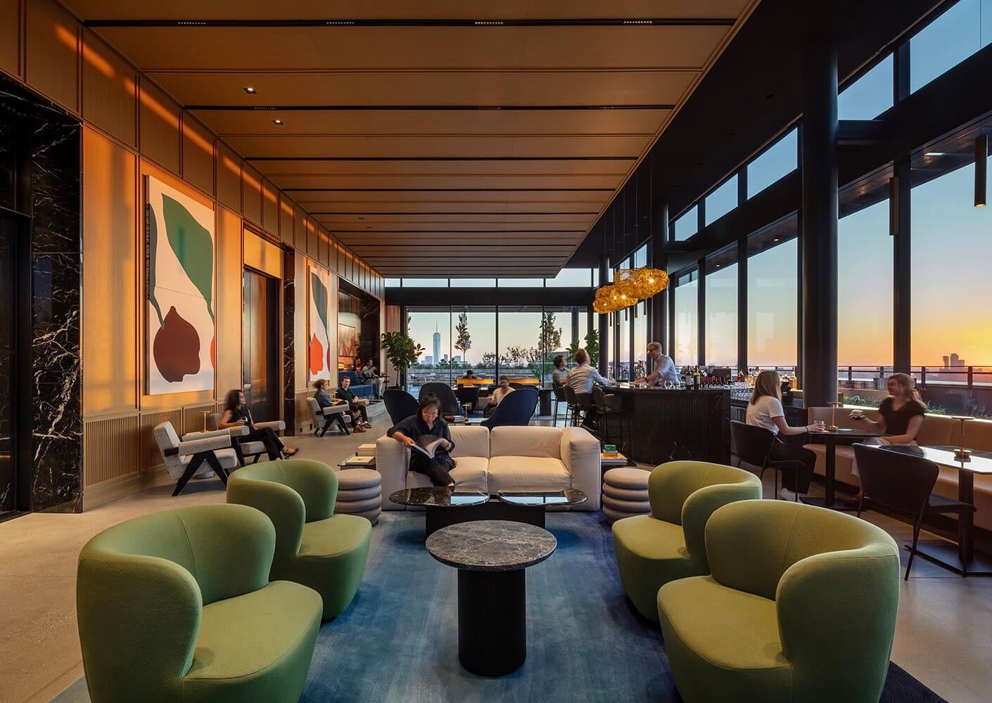 Modern lounge with green chairs, people socializing at sunset near large windows.